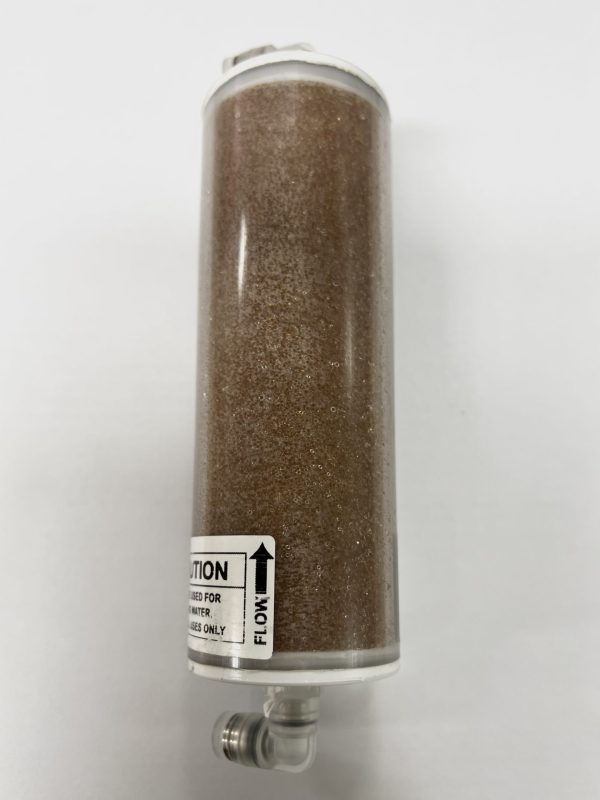 A cylindrical water filter filled with brown granules, positioned upright. It has a small attached nozzle. The label reads: "CAUTION: Used for water. Use only. FLOW ↑". Placed on a white surface.