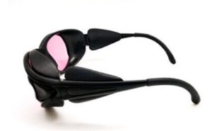 Black sunglasses with pink lenses, featuring a sleek, futuristic design. They are resting on a white surface, folded neatly with curved arms and thick frames.